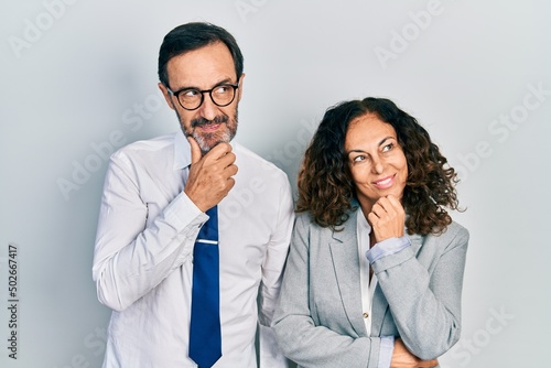 Middle age couple of hispanic woman and man wearing business office uniform with hand on chin thinking about question, pensive expression. smiling with thoughtful face. doubt concept.
