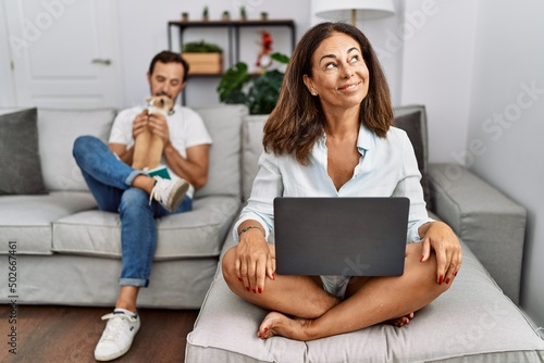 Hispanic middle age couple at home, woman using laptop smiling looking to the side and staring away thinking.