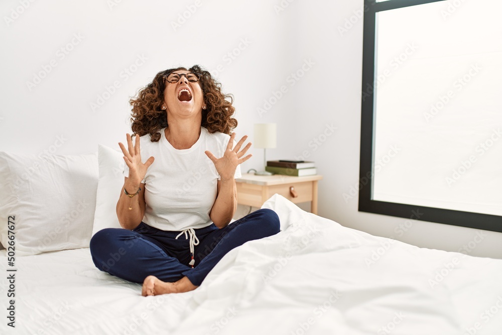 Middle age hispanic woman sitting on the bed at home crazy and mad shouting and yelling with aggressive expression and arms raised. frustration concept. Middle age hispanic woman sitting on the bed at home crazy and mad shouting and yelling with aggressive expression and arms raised. frustration concept.