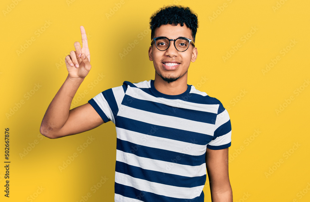 Young african american man wearing casual clothes and glasses showing and pointing up with finger number one while smiling confident and happy. Young african american man wearing casual clothes and glasses showing and pointing up with finger number one while smiling confident and happy.