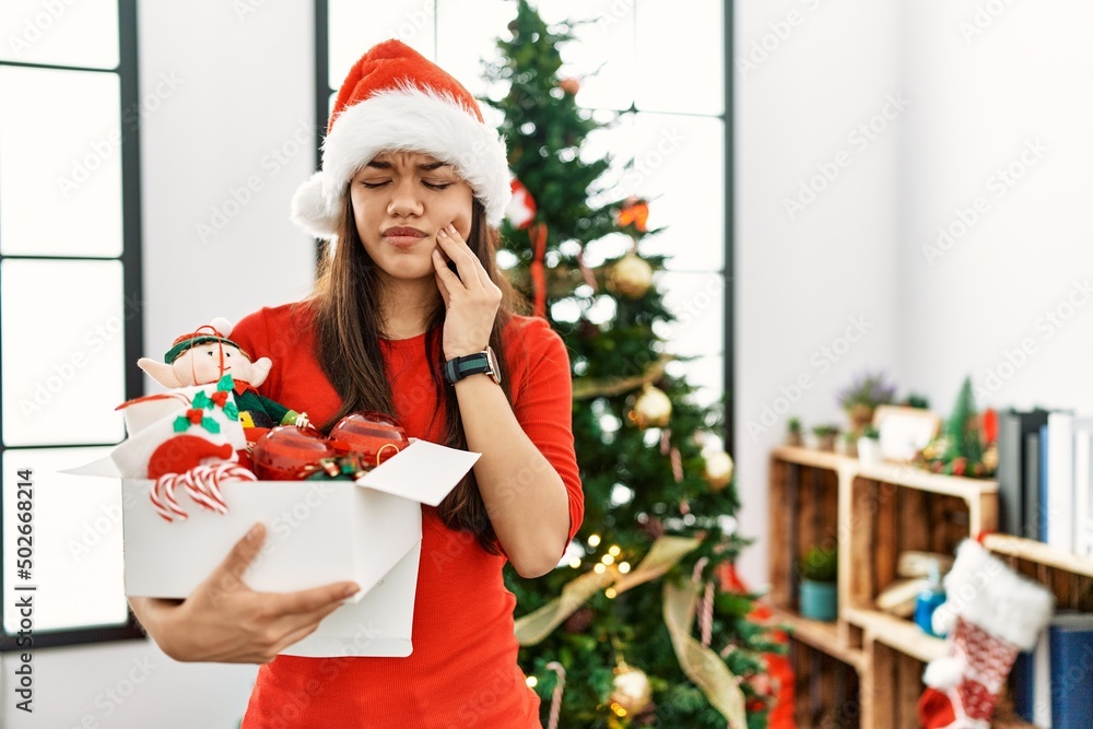 Young brunette woman standing by christmas tree holding decoration touching mouth with hand with painful expression because of toothache or dental illness on teeth. dentist concept. Young brunette woman standing by christmas tree holding decoration touching mouth with hand with painful expression because of toothache or dental illness on teeth. dentist concept.