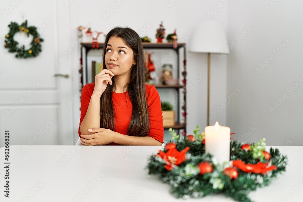 Young brunette woman sitting on the table on christmas serious face thinking about question with hand on chin, thoughtful about confusing idea Young brunette woman sitting on the table on christmas serious face thinking about question with hand on chin, thoughtful about confusing idea
