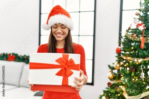 Young latin woman unboxing gift standing by christmas tree at home