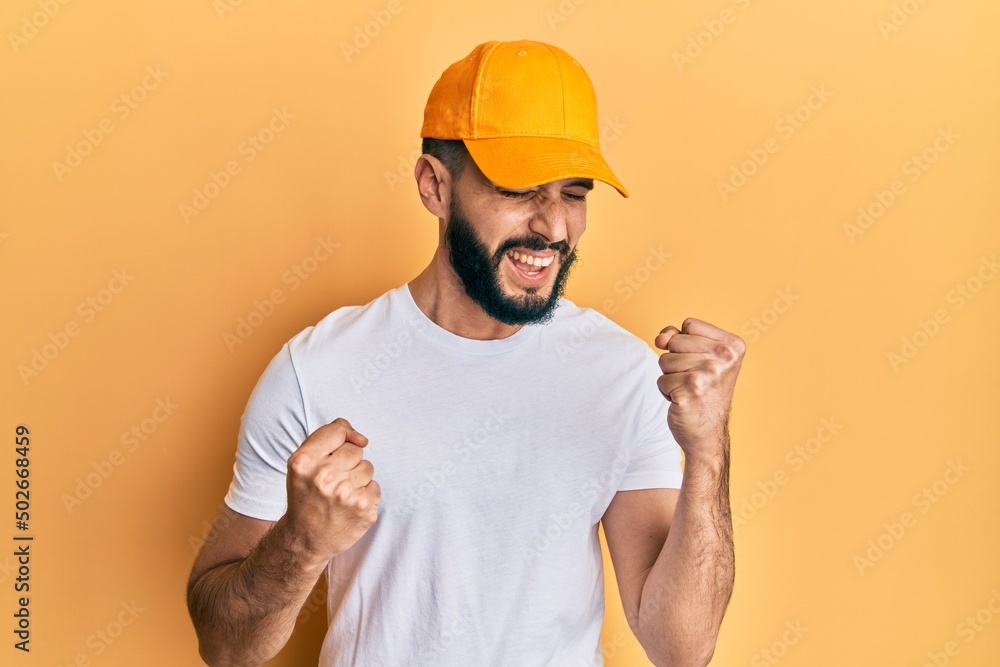 Young man with beard wearing yellow cap celebrating surprised and amazed for success with arms raised and eyes closed