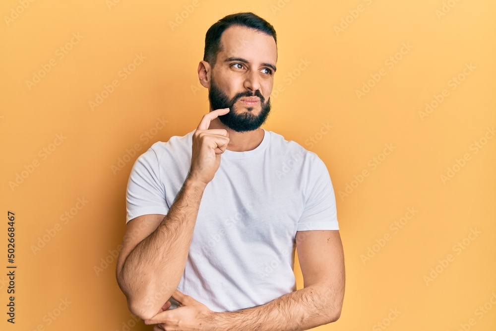 Young man with beard wearing casual white t shirt serious face thinking about question with hand on chin, thoughtful about confusing idea Young man with beard wearing casual white t shirt serious face thinking about question with hand on chin, thoughtful about confusing idea