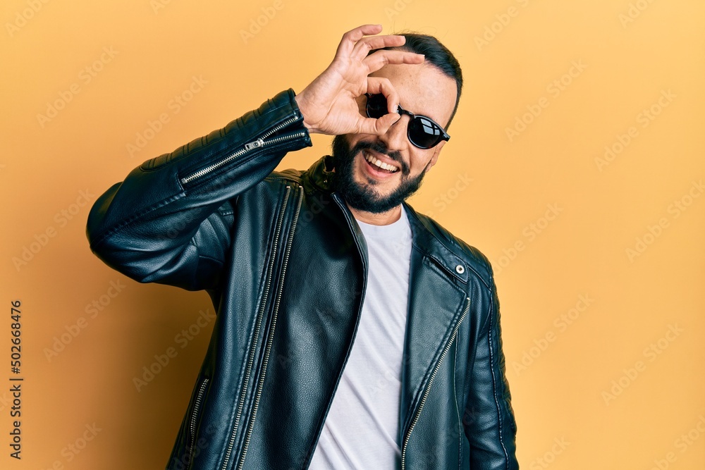 Young man with beard wearing black leather jacket and sunglasses smiling happy doing ok sign with hand on eye looking through fingers Young man with beard wearing black leather jacket and sunglasses smiling happy doing ok sign with hand on eye looking through fingers