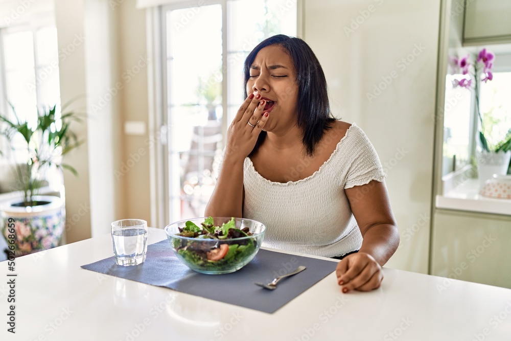 Young hispanic woman eating healthy salad at home bored yawning tired covering mouth with hand. restless and sleepiness.