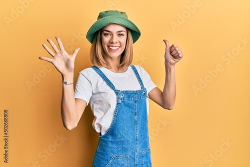 Young caucasian blonde woman wearing denim jumpsuit and hat with 90s style showing and pointing up with fingers number six while smiling confident and happy.