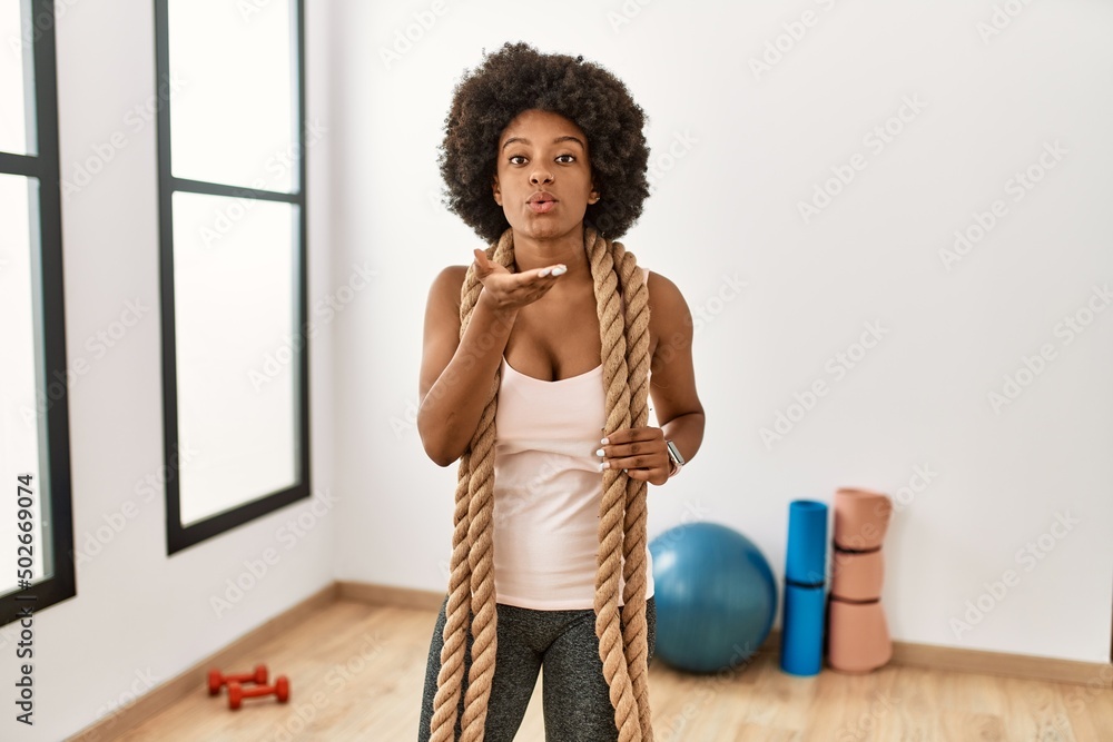 Young african american woman with afro hair at the gym training with battle ropes looking at the camera blowing a kiss with hand on air being lovely and sexy. love expression. Young african american woman with afro hair at the gym training with battle ropes looking at the camera blowing a kiss with hand on air being lovely and sexy. love expression.
