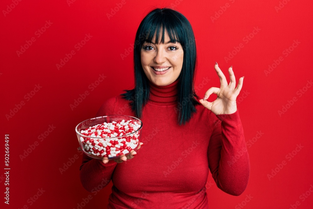 Young hispanic woman holding bowl of pills doing ok sign with fingers, smiling friendly gesturing excellent symbol Young hispanic woman holding bowl of pills doing ok sign with fingers, smiling friendly gesturing excellent symbol