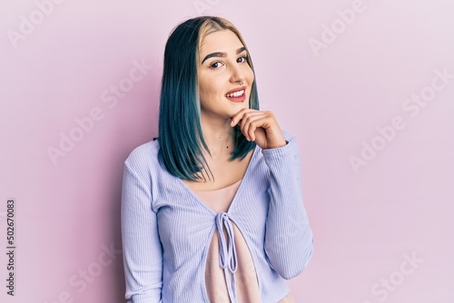 Young modern girl wearing casual jacket smiling looking confident at the camera with crossed arms and hand on chin. thinking positive.