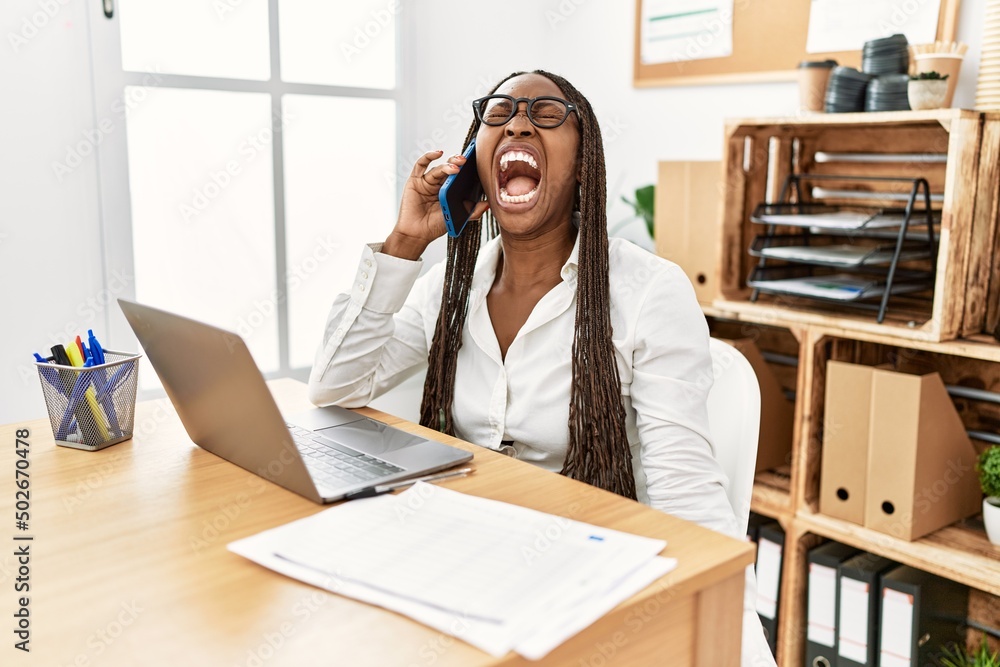 Black woman with braids working at the office speaking on the phone angry and mad screaming frustrated and furious, shouting with anger. rage and aggressive concept. Black woman with braids working at the office speaking on the phone angry and mad screaming frustrated and furious, shouting with anger. rage and aggressive concept.