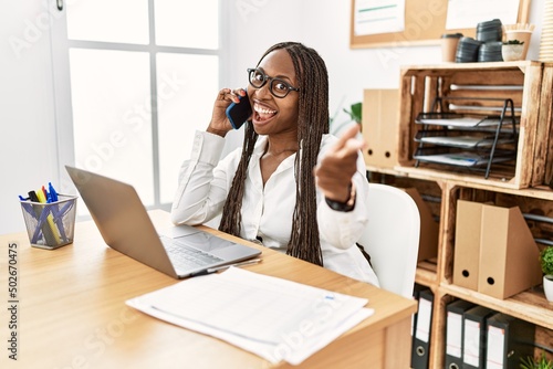 Black woman with braids working at the office speaking on the phone pointing fingers to camera with happy and funny face. good energy and vibes.