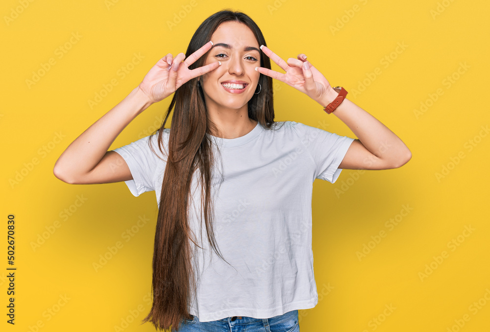 Young hispanic girl wearing casual white t shirt doing peace symbol with fingers over face, smiling cheerful showing victory Young hispanic girl wearing casual white t shirt doing peace symbol with fingers over face, smiling cheerful showing victory