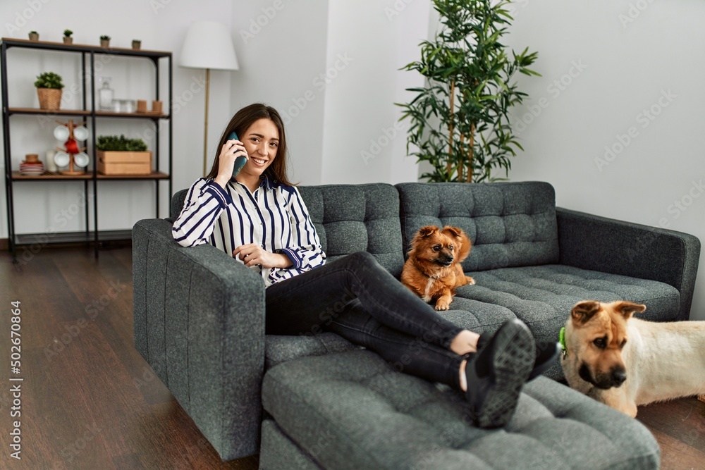 Young hispanic woman talking on the smartphone sitting on sofa with dogs at home Young hispanic woman talking on the smartphone sitting on sofa with dogs at home