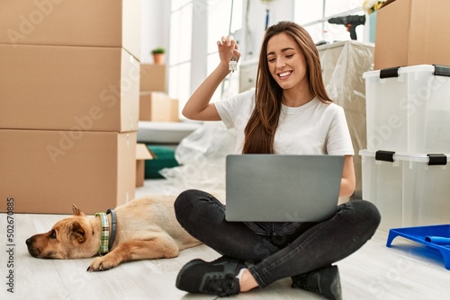 Young hispanic woman holding key having video call sitting on floor with dog at new home