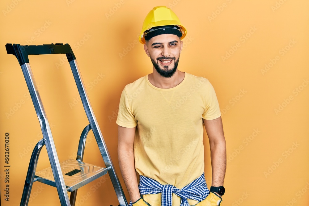 Handsome man with beard by construction stairs wearing hardhat winking looking at the camera with sexy expression, cheerful and happy face. Handsome man with beard by construction stairs wearing hardhat winking looking at the camera with sexy expression, cheerful and happy face.