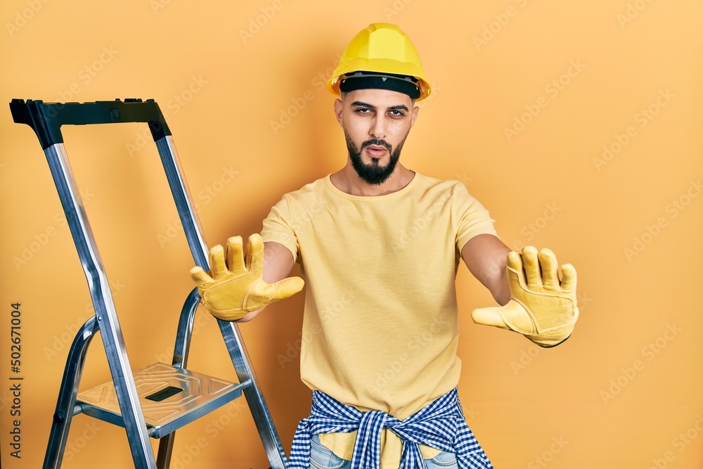 Handsome man with beard by construction stairs wearing hardhat doing stop gesture with hands palms, angry and frustration expression Handsome man with beard by construction stairs wearing hardhat doing stop gesture with hands palms, angry and frustration expression