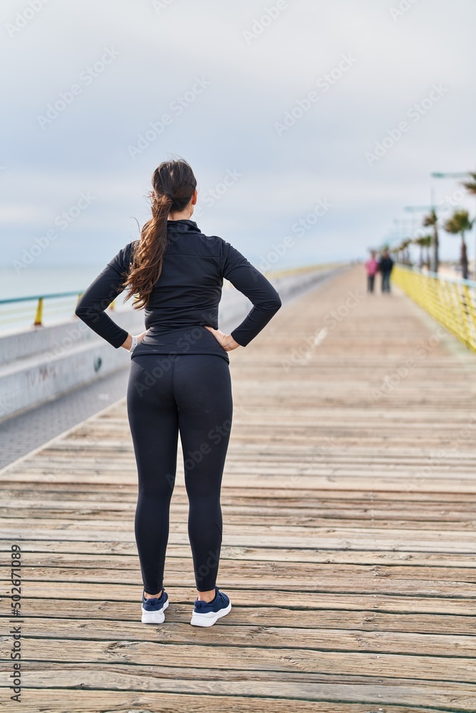 Young hispanic woman wearing sportswear standing on back view at seaside