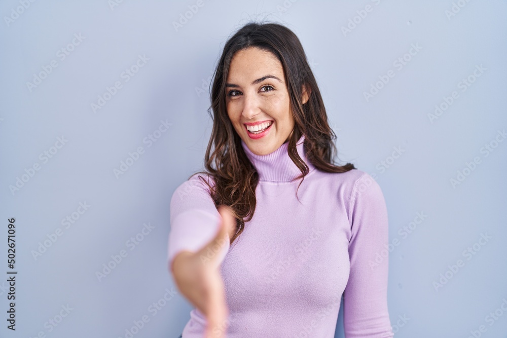 Young brunette woman standing over blue background smiling friendly offering handshake as greeting and welcoming. successful business. Young brunette woman standing over blue background smiling friendly offering handshake as greeting and welcoming. successful business.