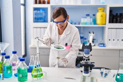 Middle age woman wearing scientist uniform looking herb with loupe at laboratory