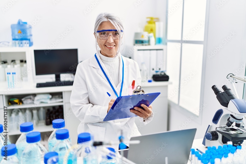 Middle age grey-haired woman wearing scientist uniform writing on clipboard working at laboratory Middle age grey-haired woman wearing scientist uniform writing on clipboard working at laboratory