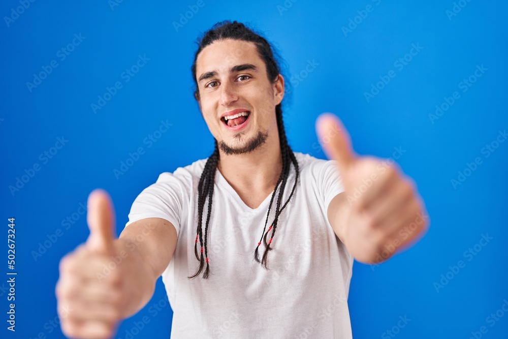 Hispanic man with long hair standing over blue background approving doing positive gesture with hand, thumbs up smiling and happy for success. winner gesture. Hispanic man with long hair standing over blue background approving doing positive gesture with hand, thumbs up smiling and happy for success. winner gesture.
