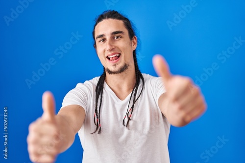 Hispanic man with long hair standing over blue background approving doing positive gesture with hand, thumbs up smiling and happy for success. winner gesture.