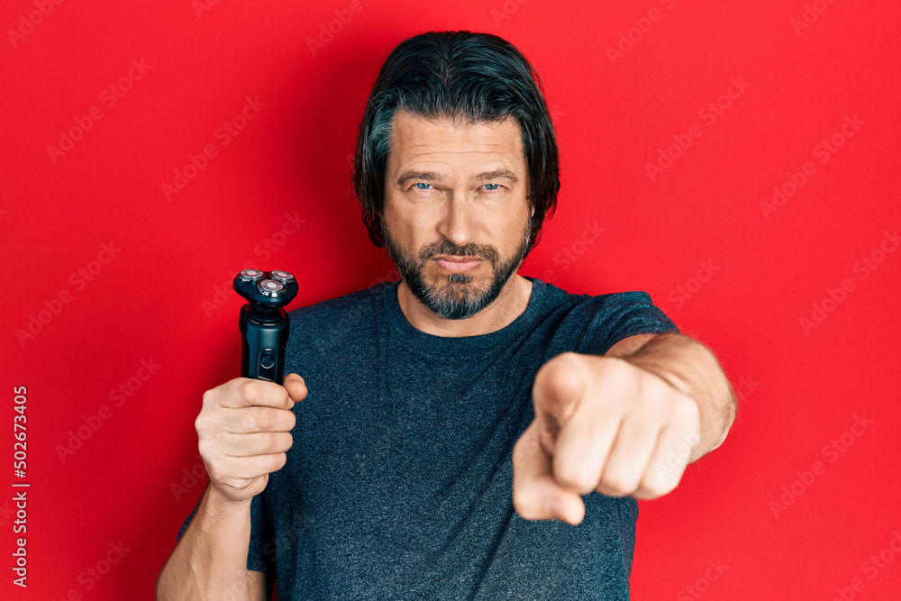 Middle age caucasian man holding electric razor machine pointing with finger to the camera and to you, confident gesture looking serious Middle age caucasian man holding electric razor machine pointing with finger to the camera and to you, confident gesture looking serious