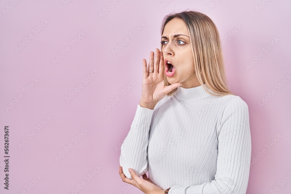 Young blonde woman wearing white sweater over pink background shouting and screaming loud to side with hand on mouth. communication concept. Young blonde woman wearing white sweater over pink background shouting and screaming loud to side with hand on mouth. communication concept.