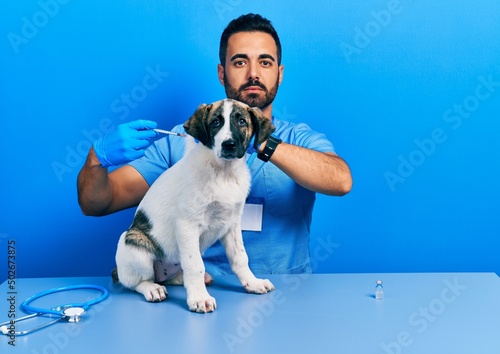 Fototapeta Naklejka Na Ścianę i Meble -  Handsome hispanic veterinary man with beard putting vaccine to puppy dog relaxed with serious expression on face. simple and natural looking at the camera.