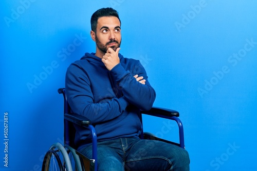 Handsome hispanic man with beard sitting on wheelchair with hand on chin thinking about question, pensive expression. smiling with thoughtful face. doubt concept.