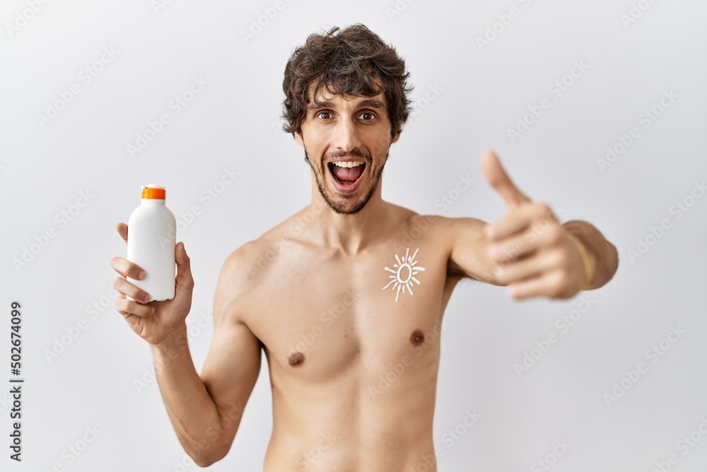 Young hispanic man standing shirtless holding sunscreen lotion approving doing positive gesture with hand, thumbs up smiling and happy for success. winner gesture.