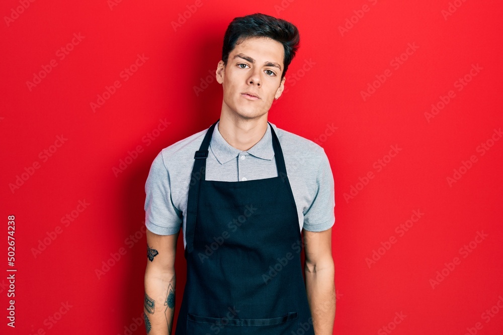 Young hispanic man wearing apron relaxed with serious expression on face. simple and natural looking at the camera. Young hispanic man wearing apron relaxed with serious expression on face. simple and natural looking at the camera.