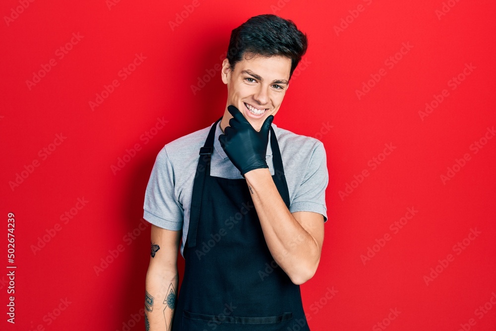 Young hispanic man wearing apron looking confident at the camera smiling with crossed arms and hand raised on chin. thinking positive. Young hispanic man wearing apron looking confident at the camera smiling with crossed arms and hand raised on chin. thinking positive.