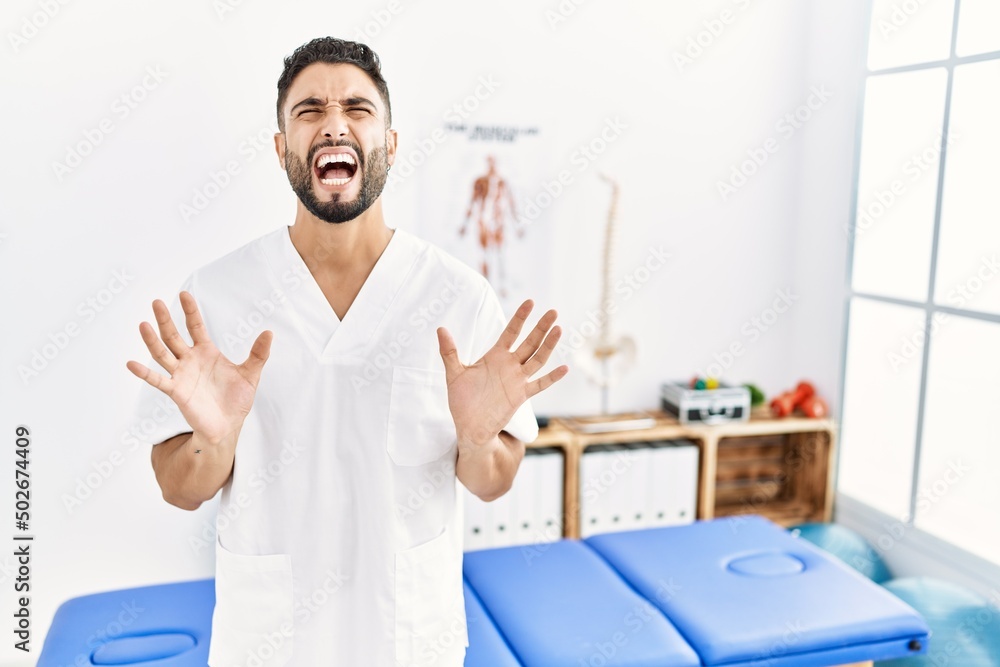 Young handsome man with beard working at pain recovery clinic crazy and mad shouting and yelling with aggressive expression and arms raised. frustration concept.
