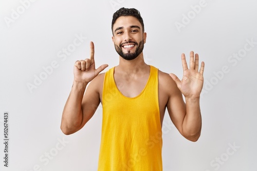 Young handsome man with beard standing over isolated background showing and pointing up with fingers number seven while smiling confident and happy.