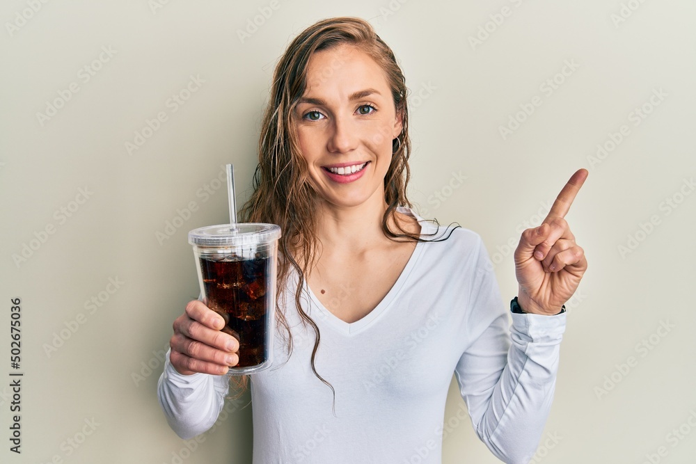 Young blonde woman drinking glass of soda beverage smiling happy pointing with hand and finger to the side Young blonde woman drinking glass of soda beverage smiling happy pointing with hand and finger to the side