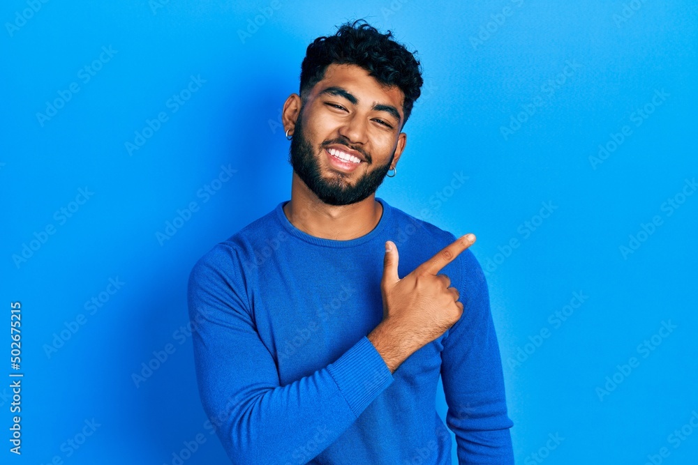 Arab man with beard wearing casual blue sweater cheerful with a smile of face pointing with hand and finger up to the side with happy and natural expression on face Arab man with beard wearing casual blue sweater cheerful with a smile of face pointing with hand and finger up to the side with happy and natural expression on face