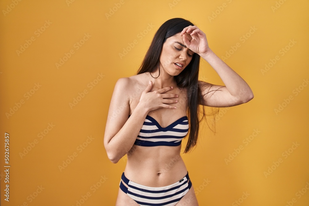 Young brunette woman wearing bikini over yellow background touching forehead for illness and fever, flu and cold, virus sick Young brunette woman wearing bikini over yellow background touching forehead for illness and fever, flu and cold, virus sick