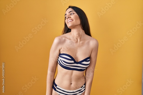 Young brunette woman wearing bikini over yellow background looking away to side with smile on face, natural expression. laughing confident.