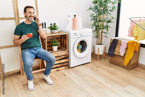 Middle age hispanic man doing laundry talking on the smartphone and drinking coffee at home.