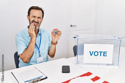 Middle age man with beard sitting by ballot holding i vote badge touching mouth with hand with painful expression because of toothache or dental illness on teeth. dentist concept.