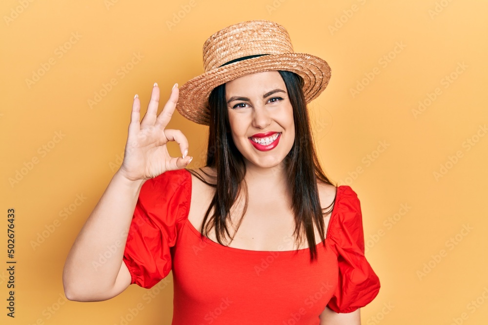 Young hispanic woman wearing summer hat smiling positive doing ok sign with hand and fingers. successful expression. Young hispanic woman wearing summer hat smiling positive doing ok sign with hand and fingers. successful expression.
