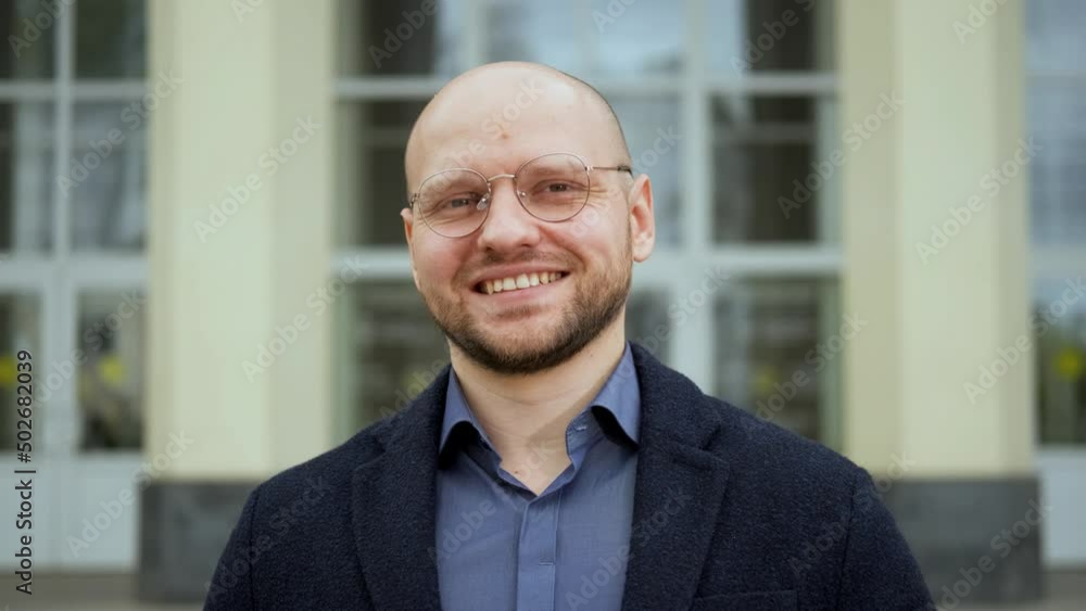 A young man, an intelligent professor, stands against the background of the university, looks at the camera and smiles. Close-up portrait