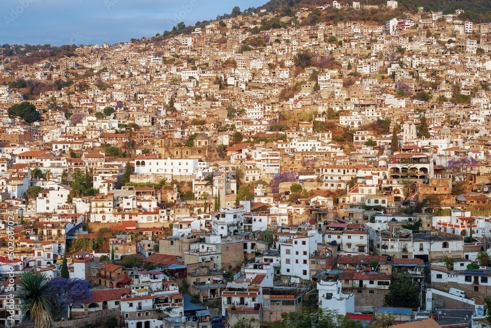 The city is illuminated by the sun in the morning on a mountainside with colonial architecture, many white houses with tiles. The cramped streets of Taxco in Mexico