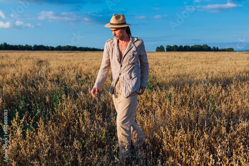 Tall handsome man dressed in a coarse linen suit and hat standing at golden oat field © Smile