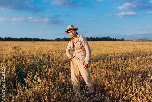 Tall handsome man dressed in a coarse linen suit and hat standing at golden oat field © Smile