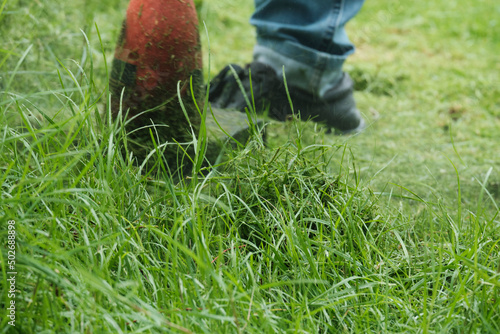 Close-up shot of male worker in jeans maintenance garden using an electric lawnmower to trim overgrown green grass in the lawn yard, scattering the grass in summer, outdoor landscaping work service.