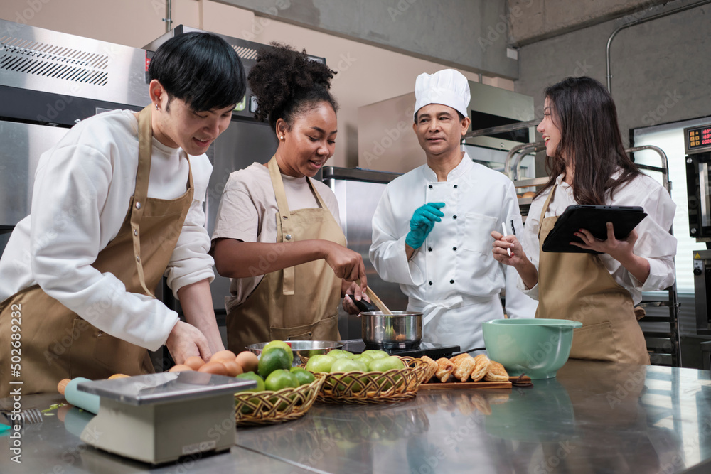 Hobby cuisine course, senior male chef in cook uniform teaches young cooking class students to prepare, mix and stir ingredients for pastry foods, fruit pies in restaurant stainless steel kitchen. Hobby cuisine course, senior male chef in cook uniform teaches young cooking class students to prepare, mix and stir ingredients for pastry foods, fruit pies in restaurant stainless steel kitchen.